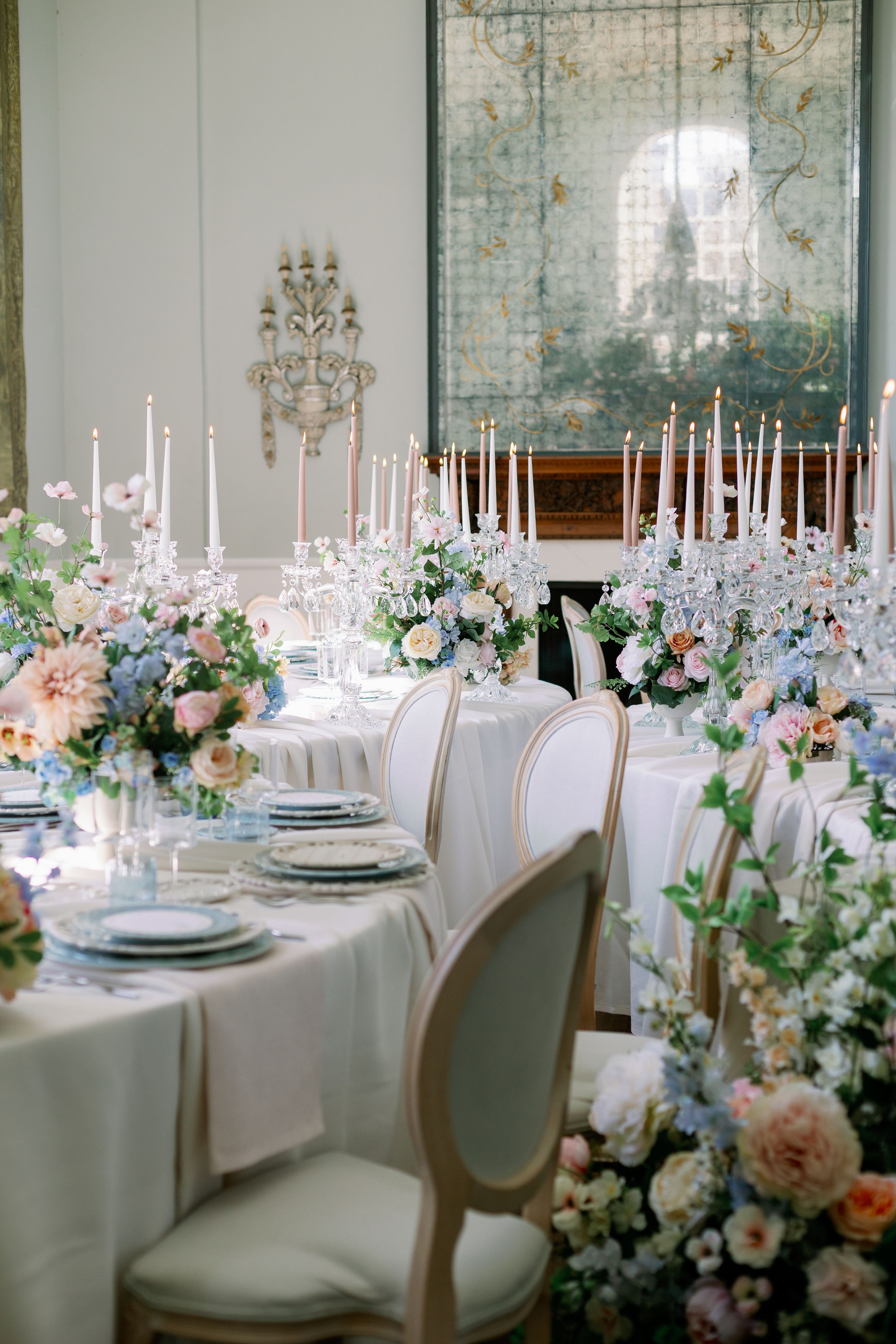 Elegant wedding reception table setting with tall white candles, pastel floral arrangements and vintage-style chairs.