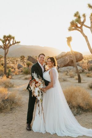 A wedding couple poses in front of Joshua trees during golden hour sunset in a desert landscape.