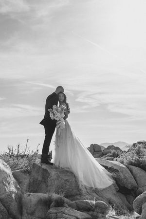 A romantic wedding couple stands atop rocky terrain in black and white as their dress flows in the desert breeze.