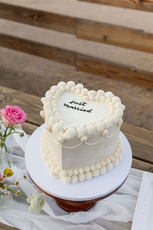 Heart-shaped white frosted cake with delicate piped border and pink rose decoration displayed on rustic wooden table.