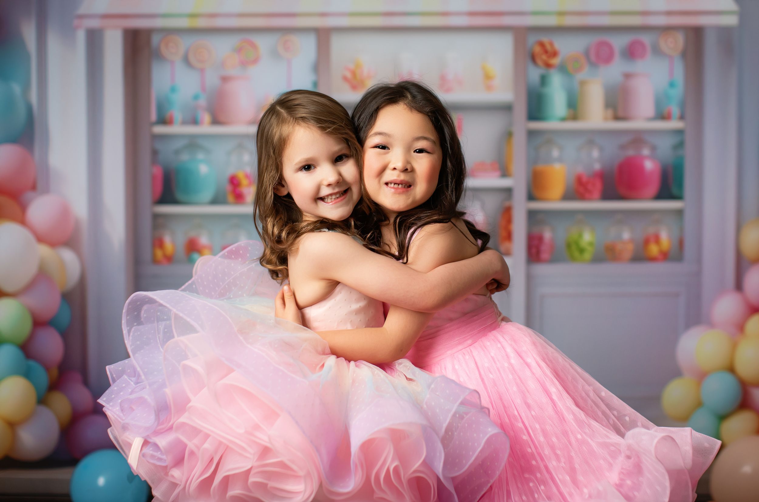 Sweet portrait of two young dancers in pink tutus hugging against candy shop themed backdrop.