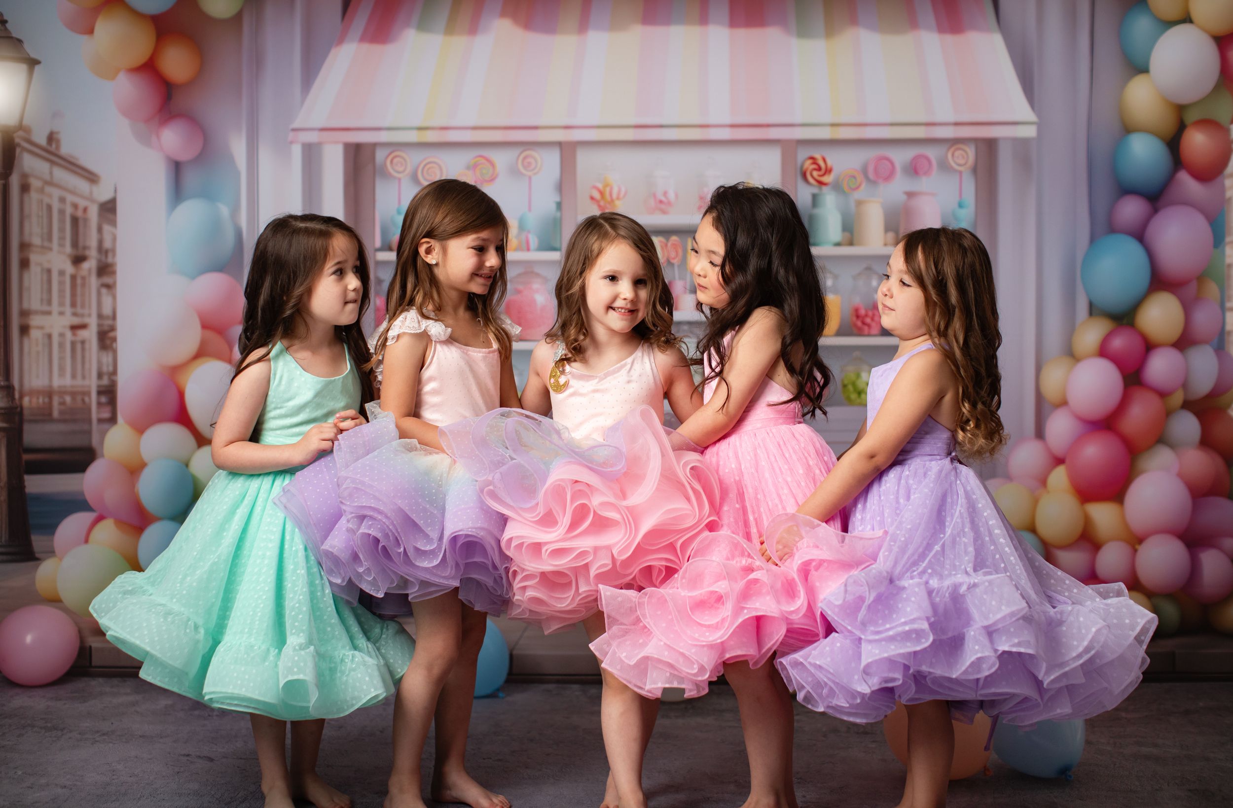Group of dancers in pastel tutus posing together against candy shop themed backdrop with balloon arch.