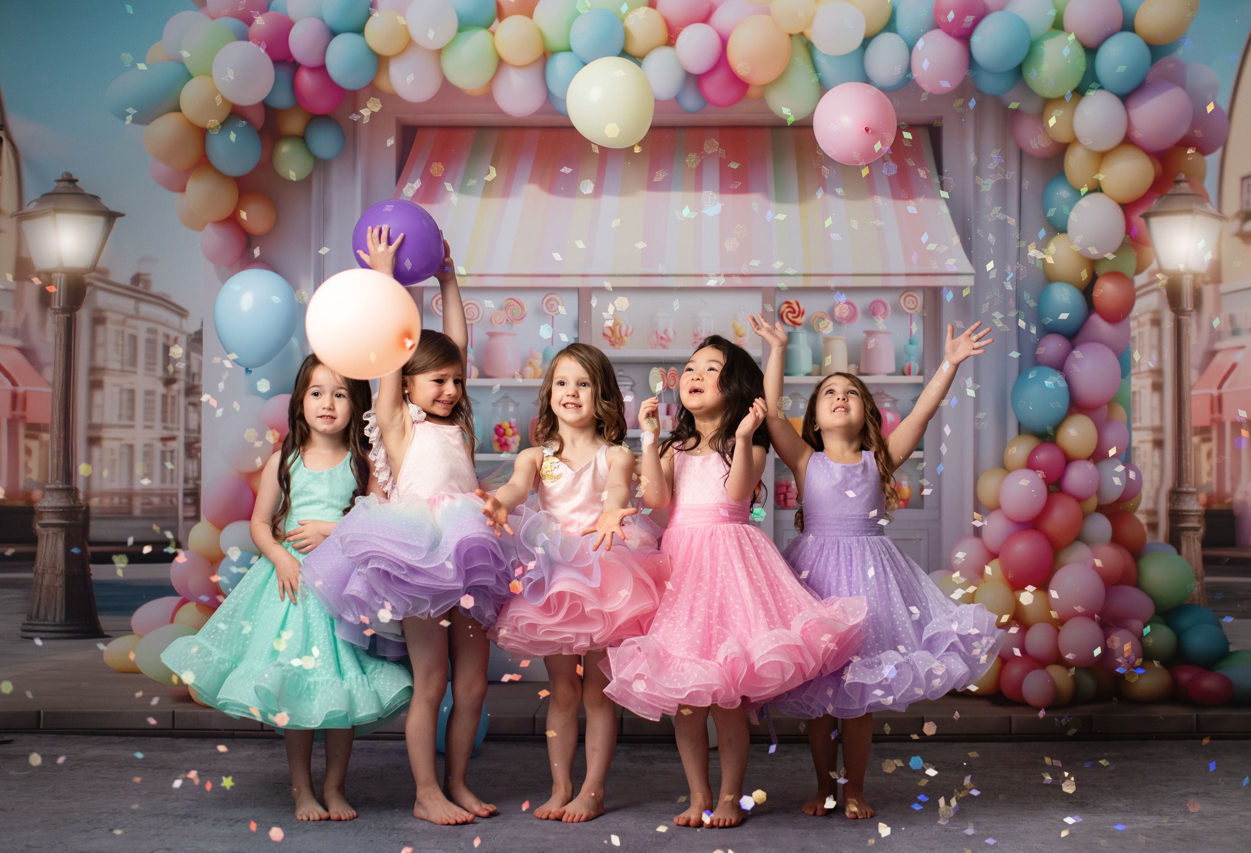 Dancing children in colorful tutus against pastel balloon arch and candy shop themed photography backdrop.