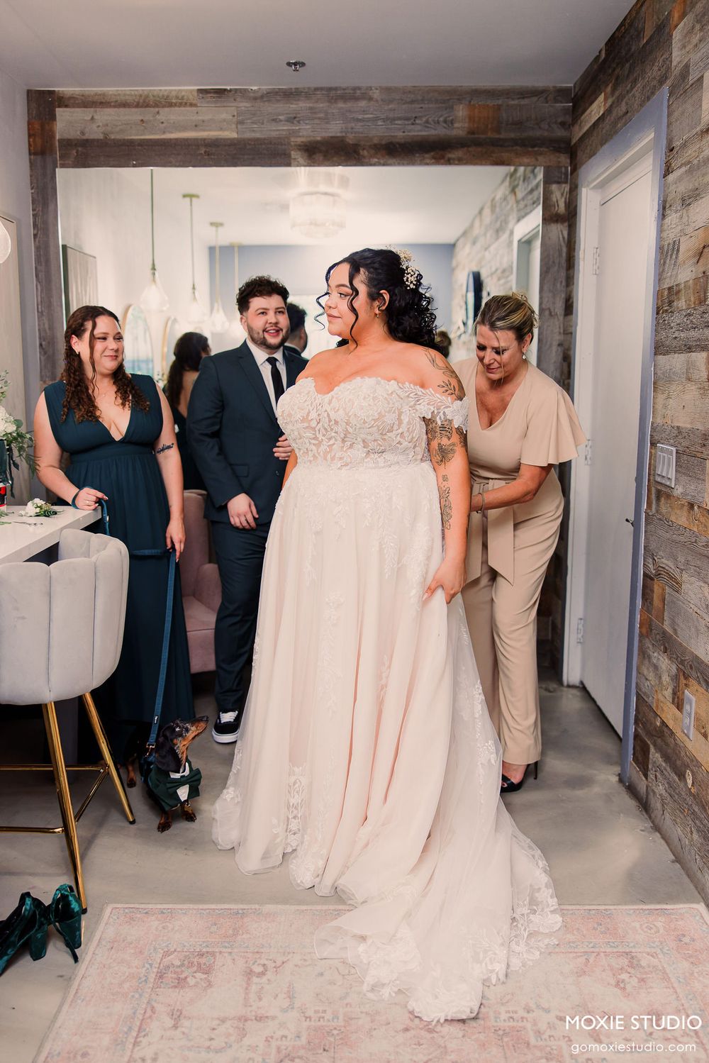 Wedding party members in blush pink dresses and tan suits gather in a rustic indoor venue.