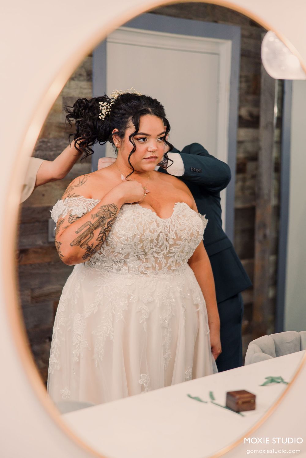 A bride in an elegant off-shoulder lace wedding gown poses gracefully in an indoor setting.
