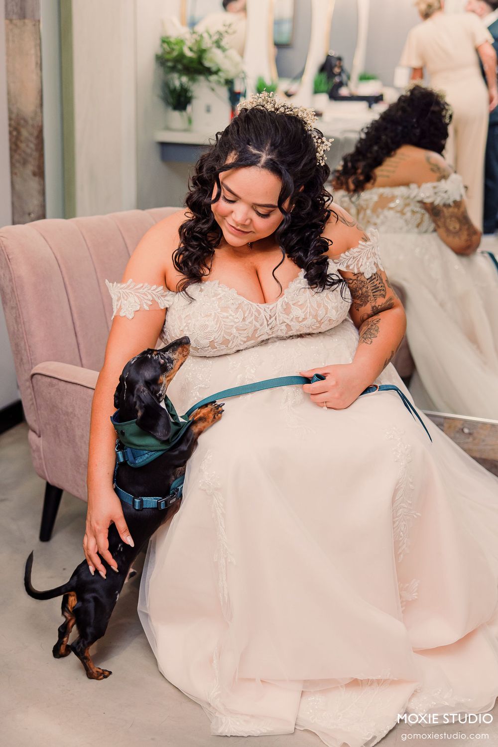 A bride in a flowing white dress sits on a plush chair while interacting with a black service dog.