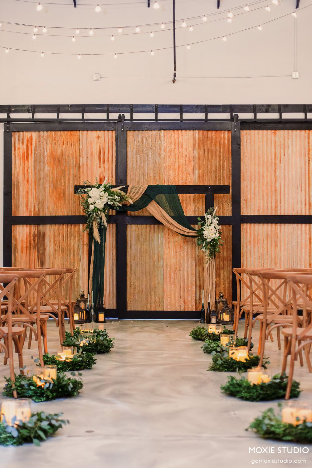 String lights illuminate a rustic wedding ceremony setup with wooden cross-back chairs and greenery floor arrangements.