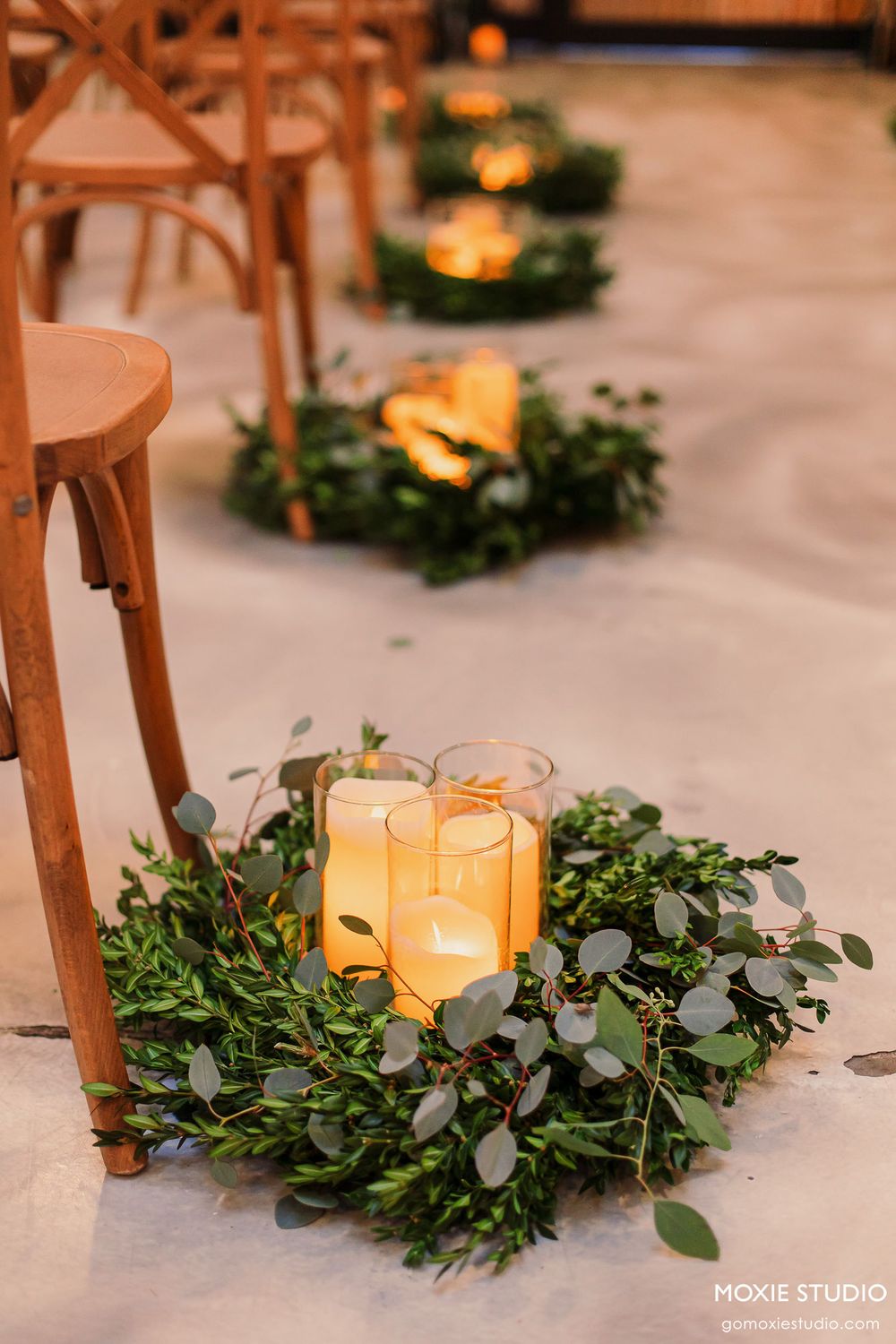 Rustic wedding decor featuring glowing candles surrounded by fresh eucalyptus greenery garlands on white tablecloths.