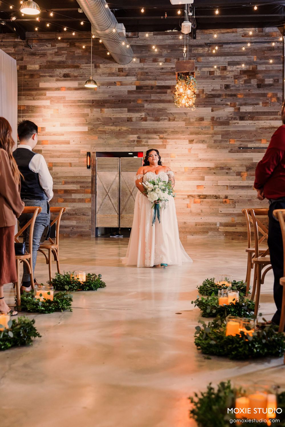 Bride in white gown holding bouquet stands in rustic venue with wooden walls and string lights overhead.