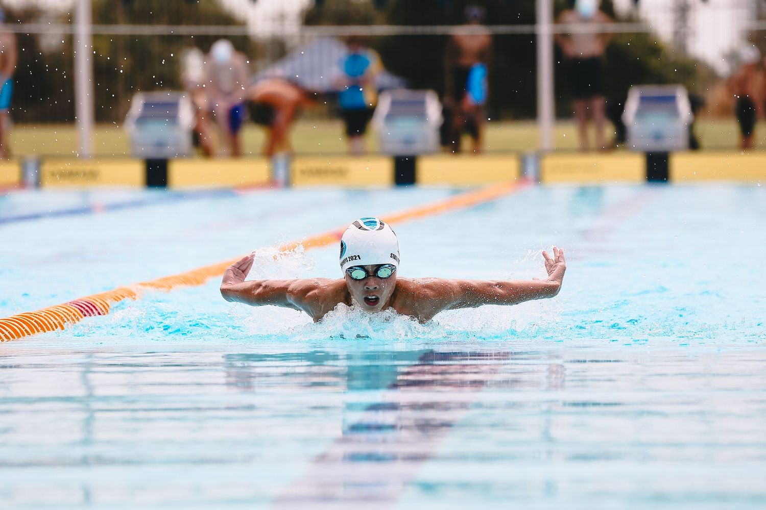 Swimmer executes butterfly stroke with outstretched arms in outdoor competition pool.