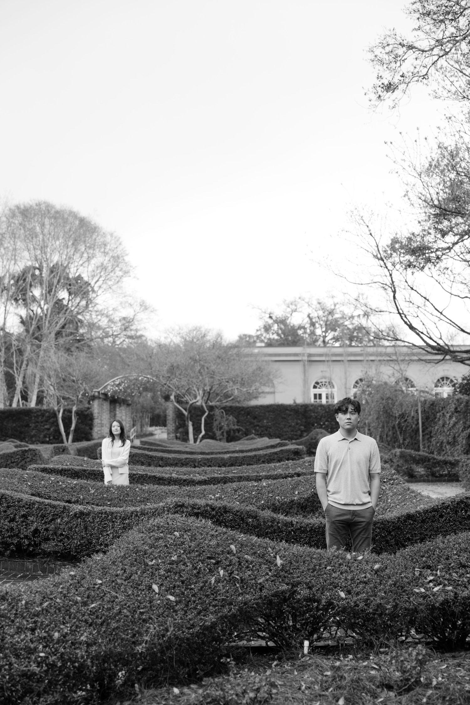 Black and white photo of person standing in formal garden with sculptured hedges and classical architecture in background.