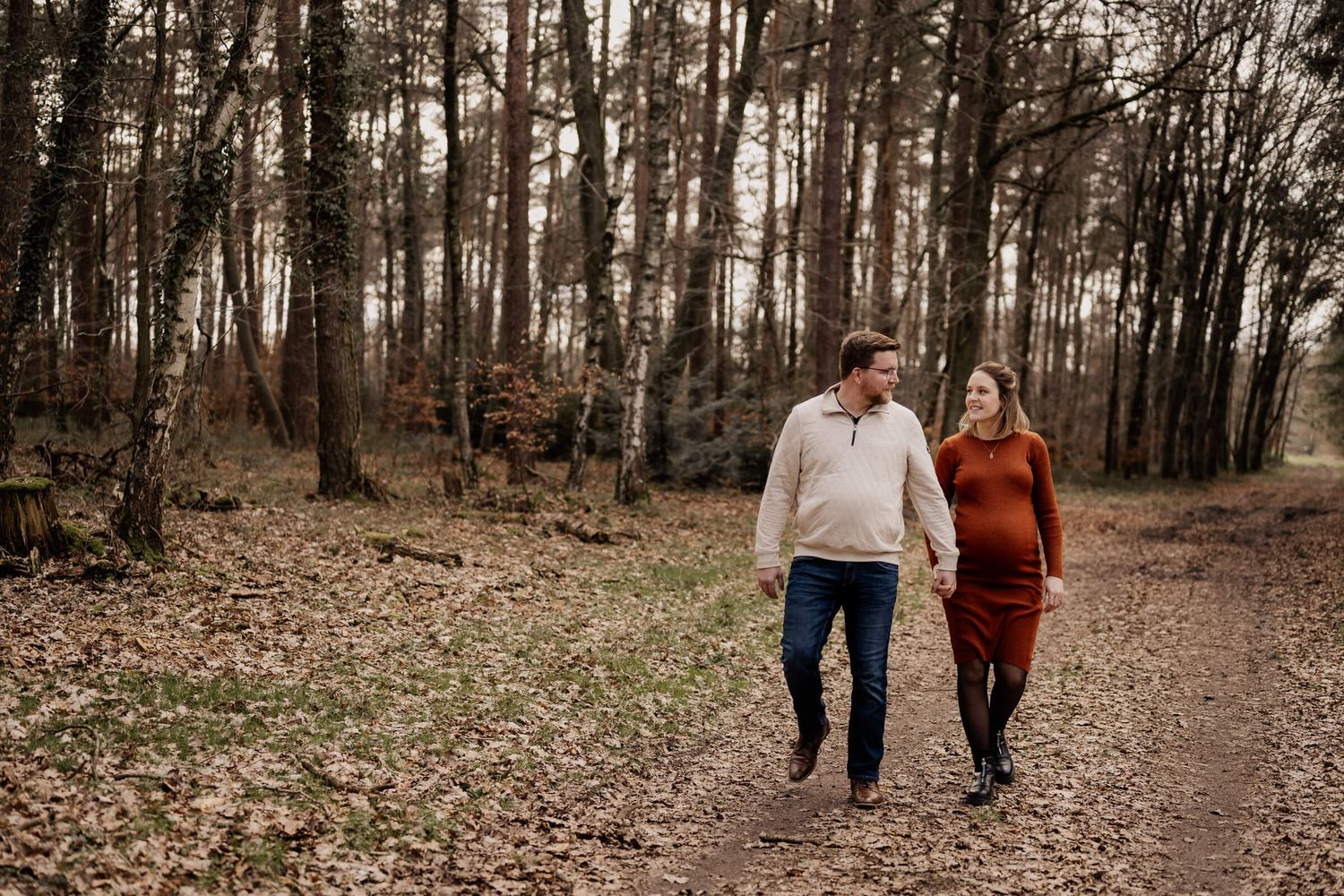 A couple walks hand in hand along a forest path during autumn, surrounded by tall trees and fallen leaves.