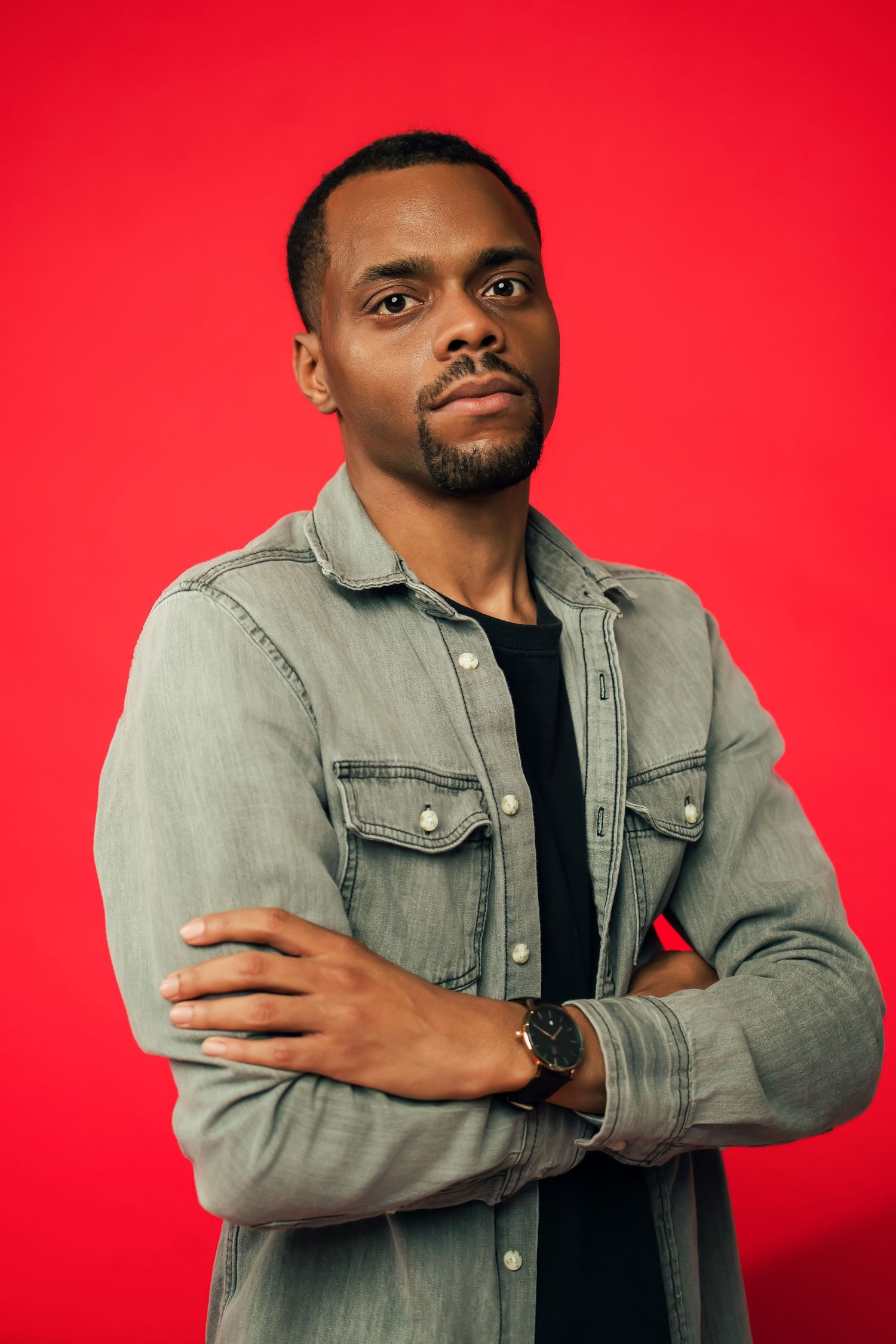 Portrait of a person wearing a denim jacket and black t-shirt posing confidently against a bright red background.