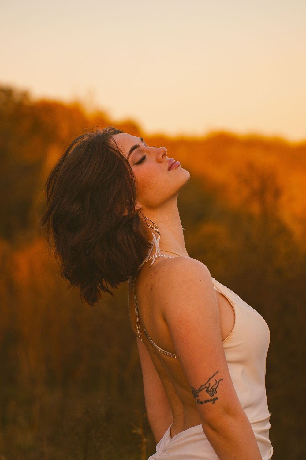 Woman in white top poses against golden sunset backdrop with dramatic lighting.