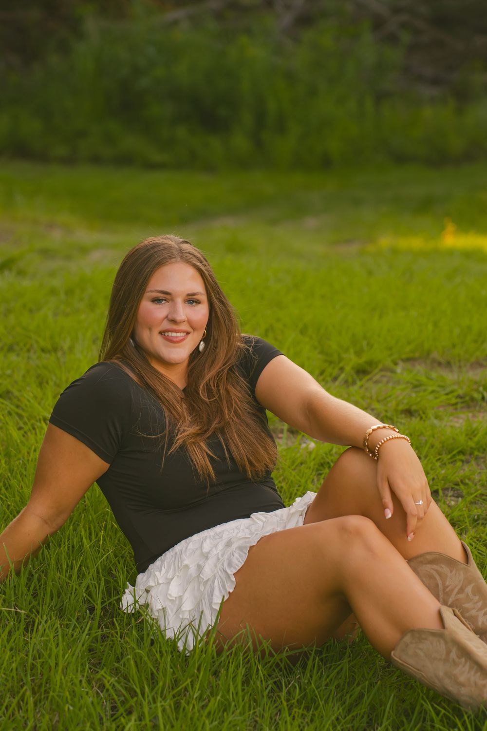 Woman in black top and white skirt relaxing in green grass field during outdoor photoshoot.