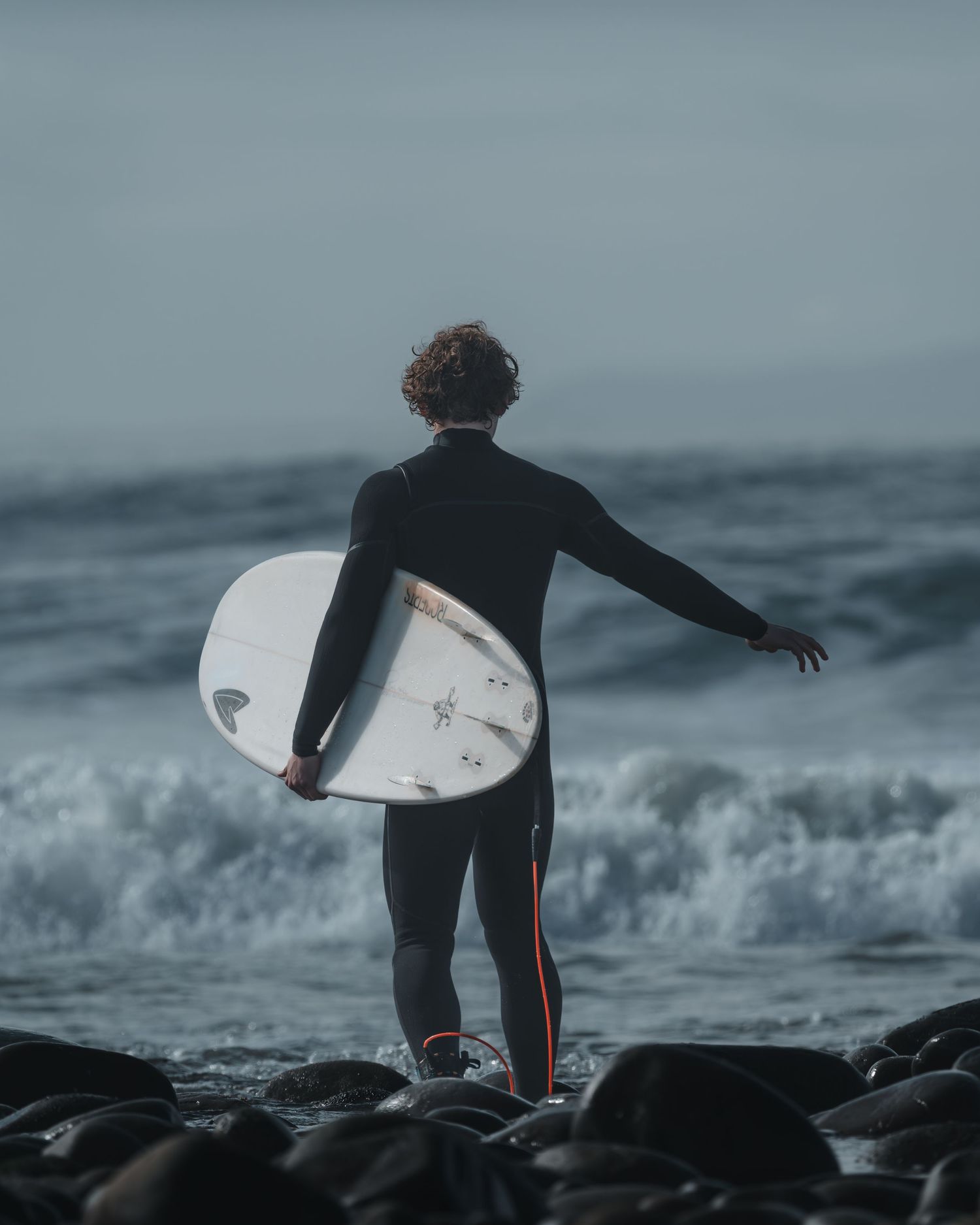 Silhouetted surfer stands on rocky beach holding surfboard against stormy ocean waves.