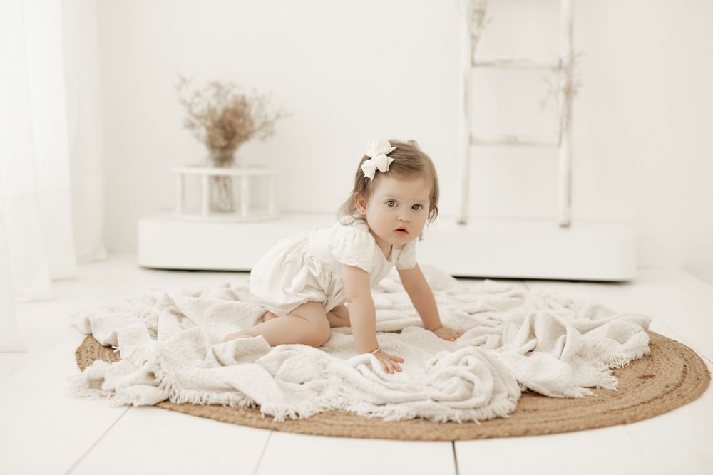 Baby in white lace dress crawling on round jute rug with soft neutral decor in background.