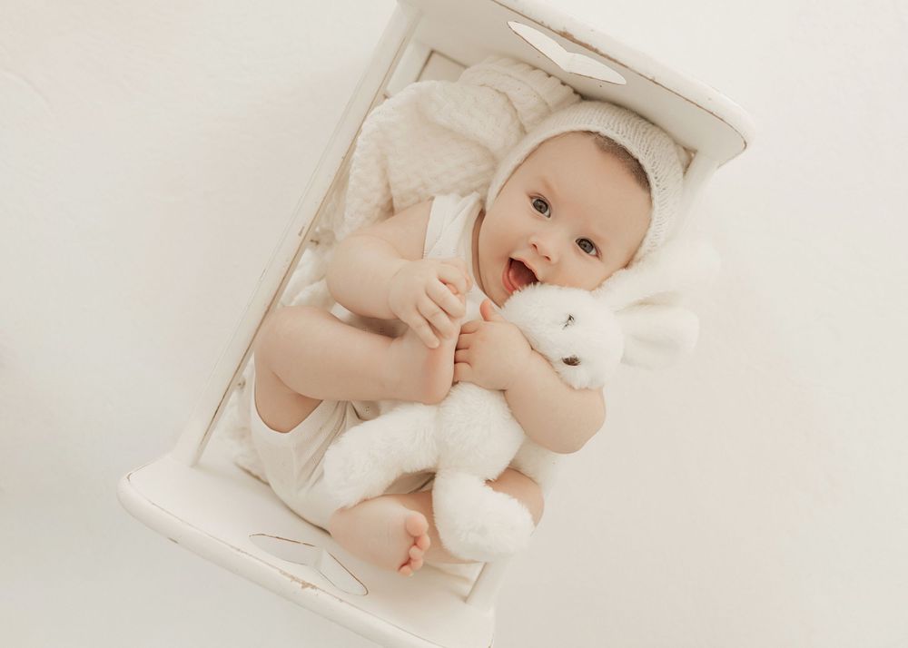 Baby in a cream-colored onesie lying on white bedding while holding a small plush teddy bear toy.