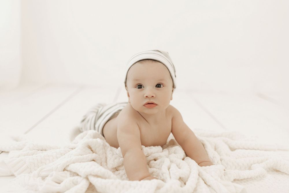 A baby wearing a white hat lies on soft white blankets in a minimalist studio setting.