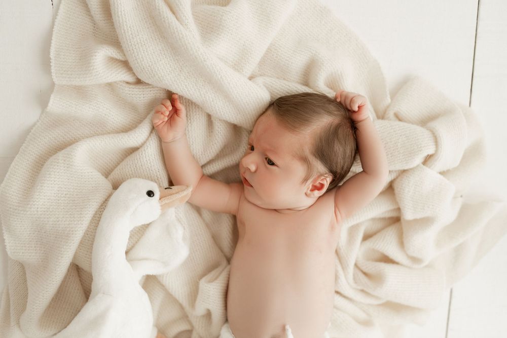 A sleeping infant lies on a cream blanket next to a white plush duck toy.