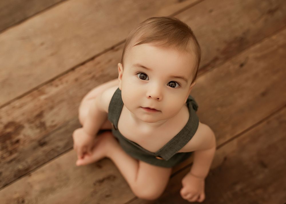 Baby wearing olive green top sits on rustic wooden floor during photo session.
