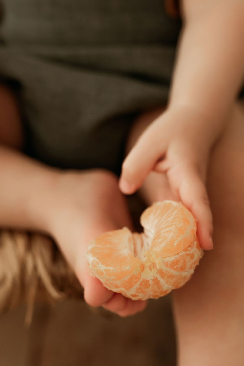 Small hands hold a peeled orange segment showing the juicy citrus fruit interior against a dark background.