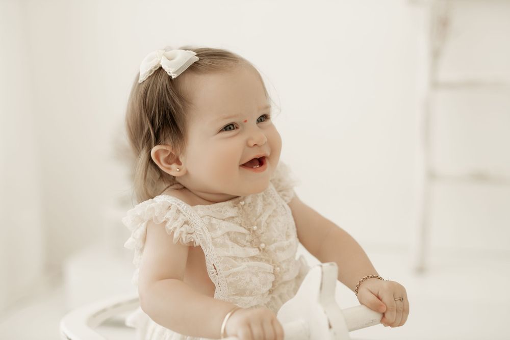 A smiling baby wearing a white lace dress and small bow sits while posing for a portrait in soft lighting.
