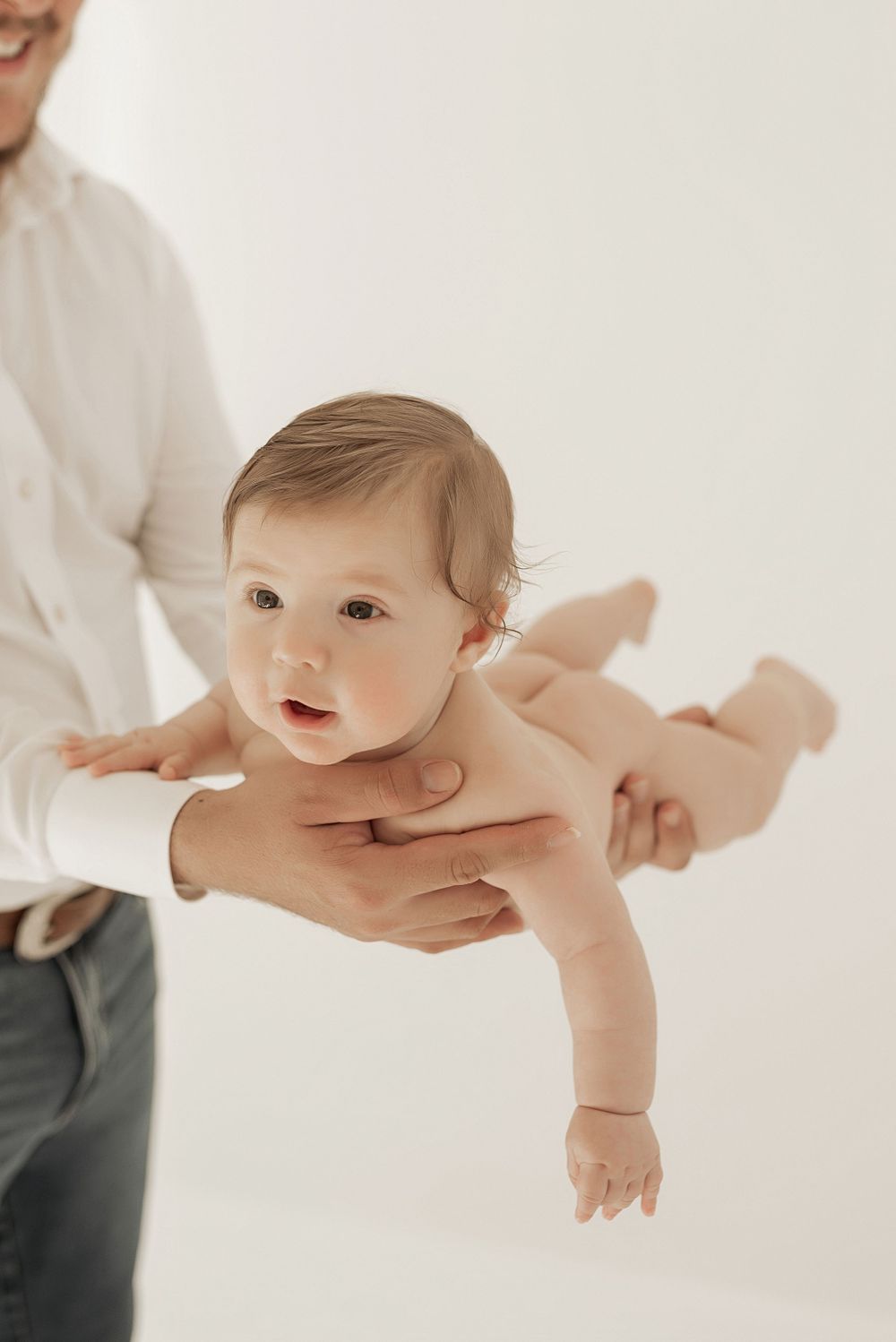 Baby wearing diaper held up safely in the air by adult hands in a white studio setting.