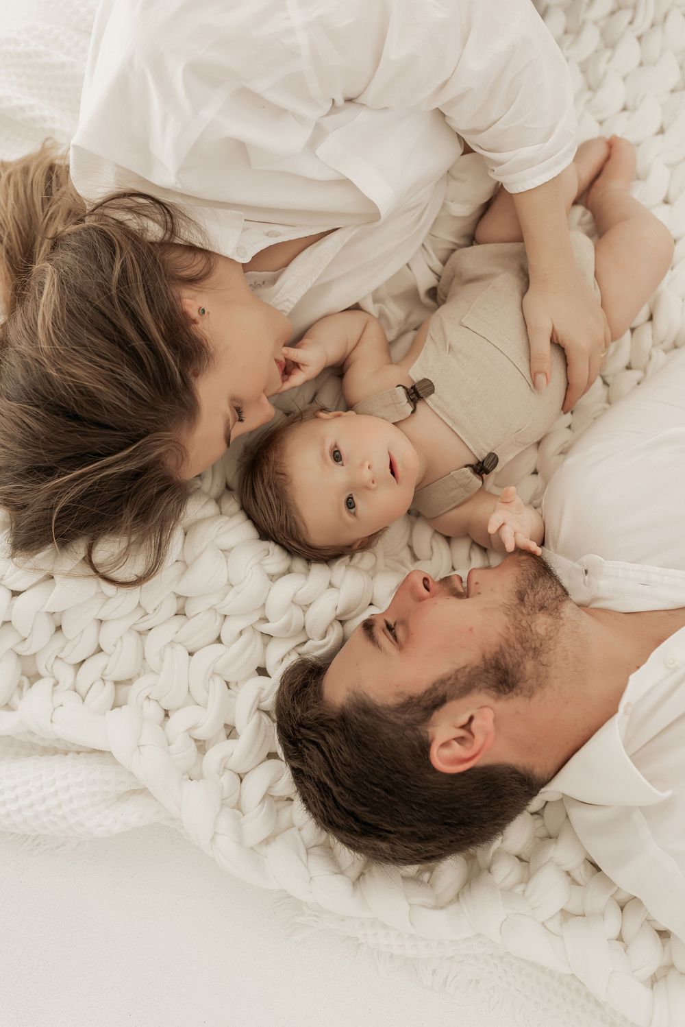 A family lying together on a cozy white knit blanket wearing matching white outfits.