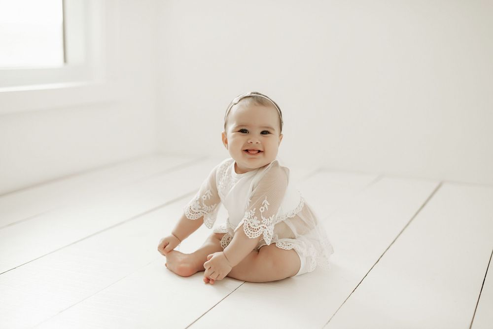 Baby wearing delicate white lace romper sits on white wooden floor in bright minimalist setting.
