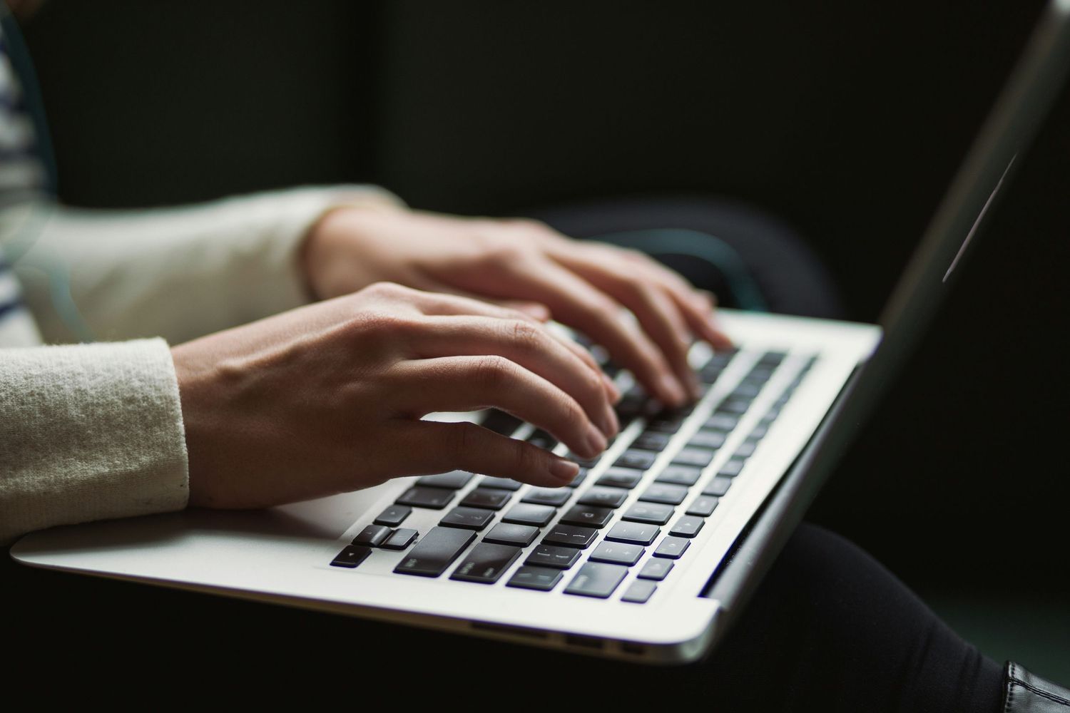 Hands typing on a silver laptop keyboard in dimly lit environment with soft focus.