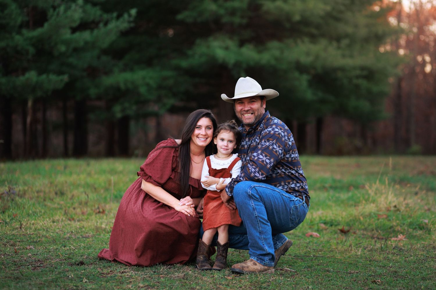 Cute country family pose in front of large pine trees in the Illinois evening sunset.