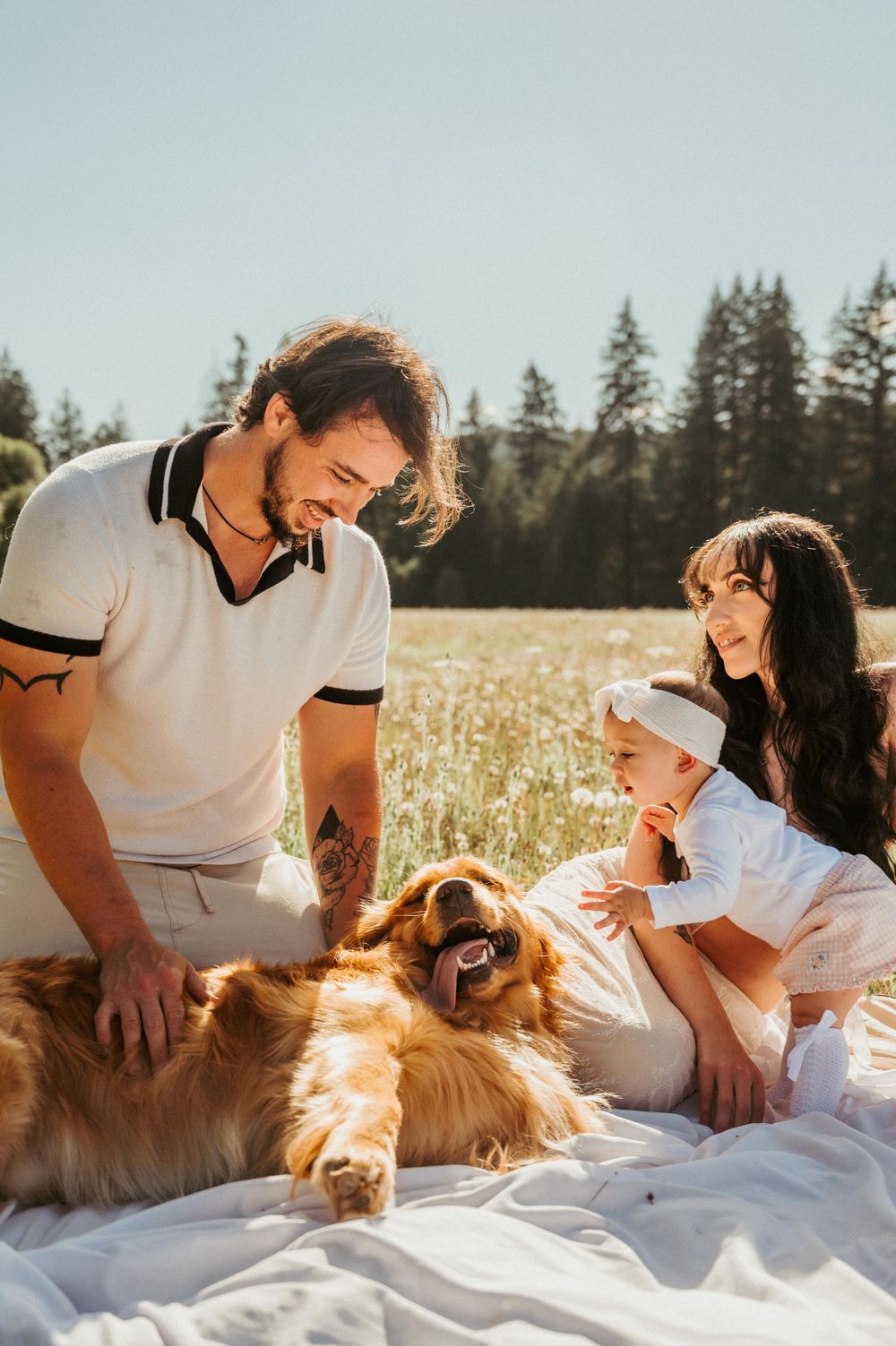 Family relaxing on white blanket with golden retriever in sunny meadow.