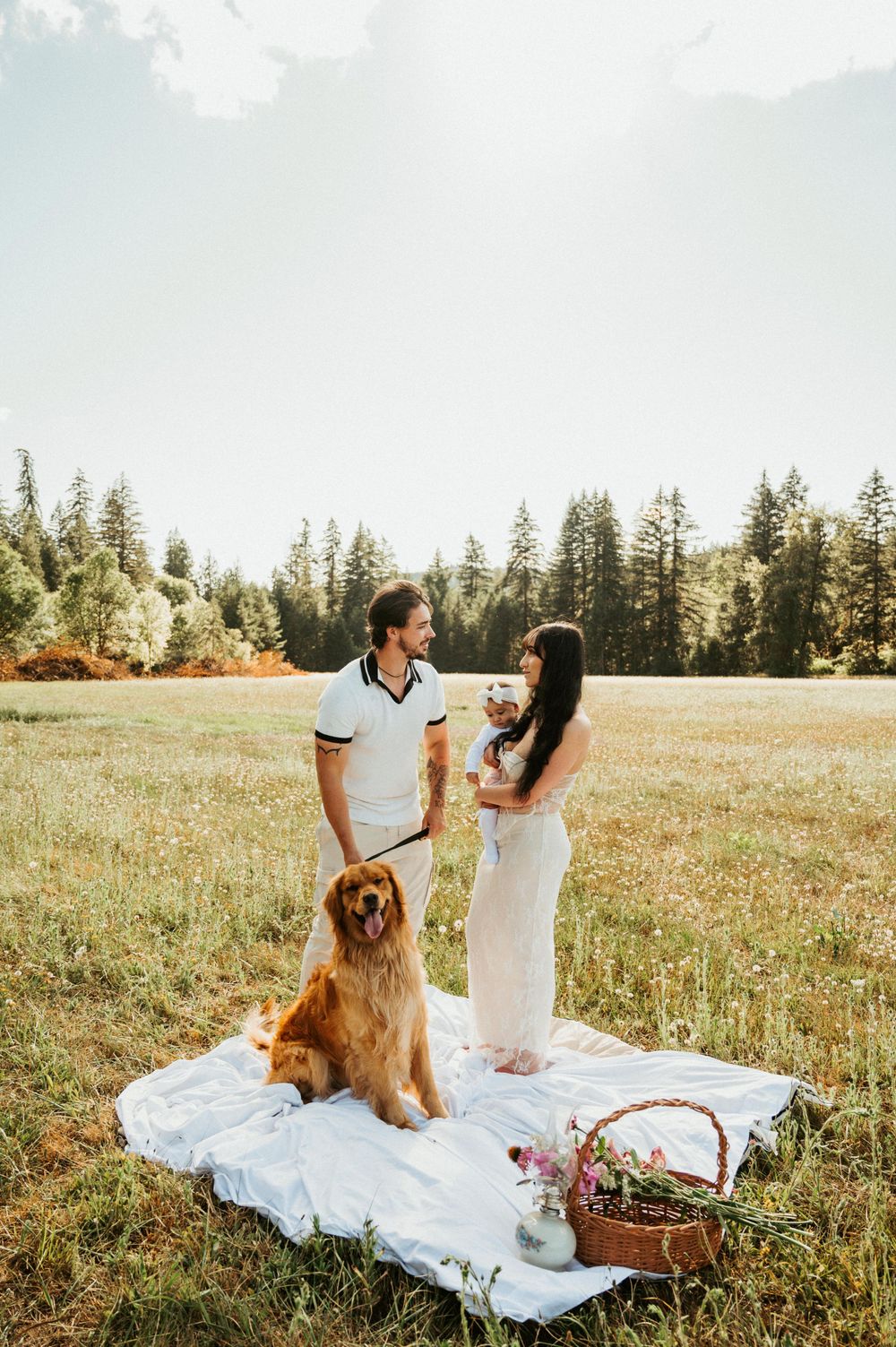 Family and golden retriever having picnic on white blanket in sunny meadow with pine trees.
