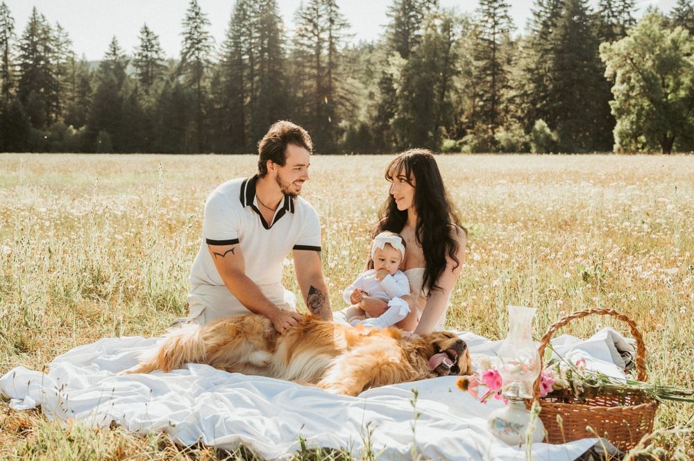 Family and golden retriever enjoying picnic on white blanket in sunny field.