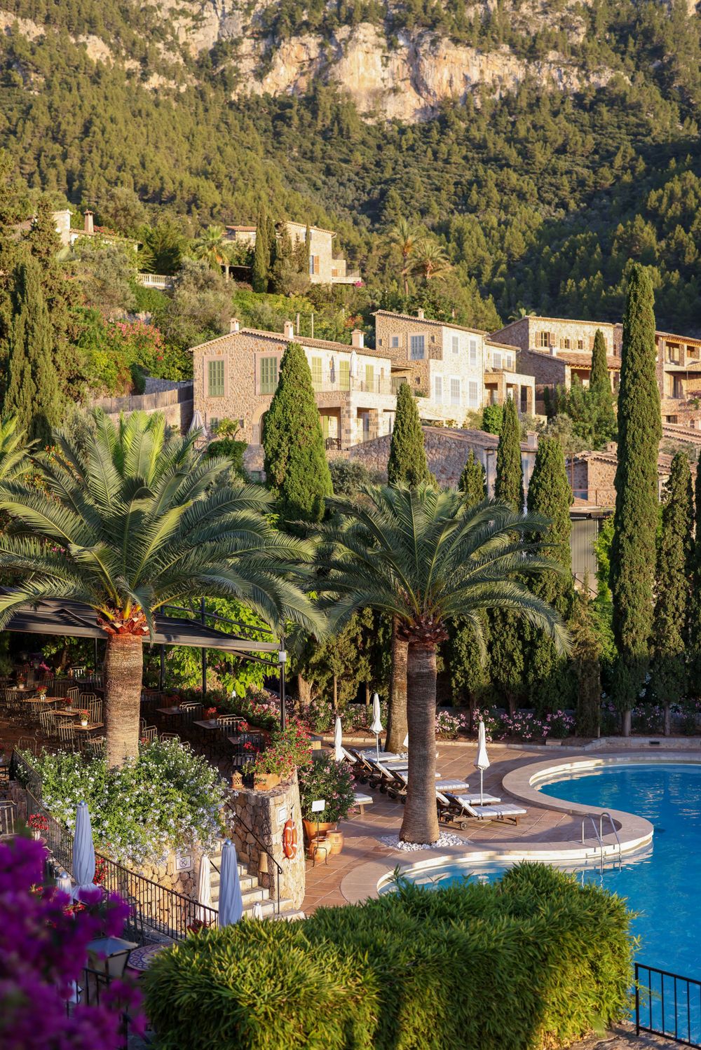 Luxury resort with curved swimming pool and palm trees against mountainous Mediterranean landscape.