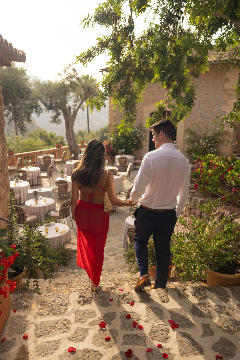 Rose petal-strewn stone pathway leading to romantic outdoor dining setup at sunset.