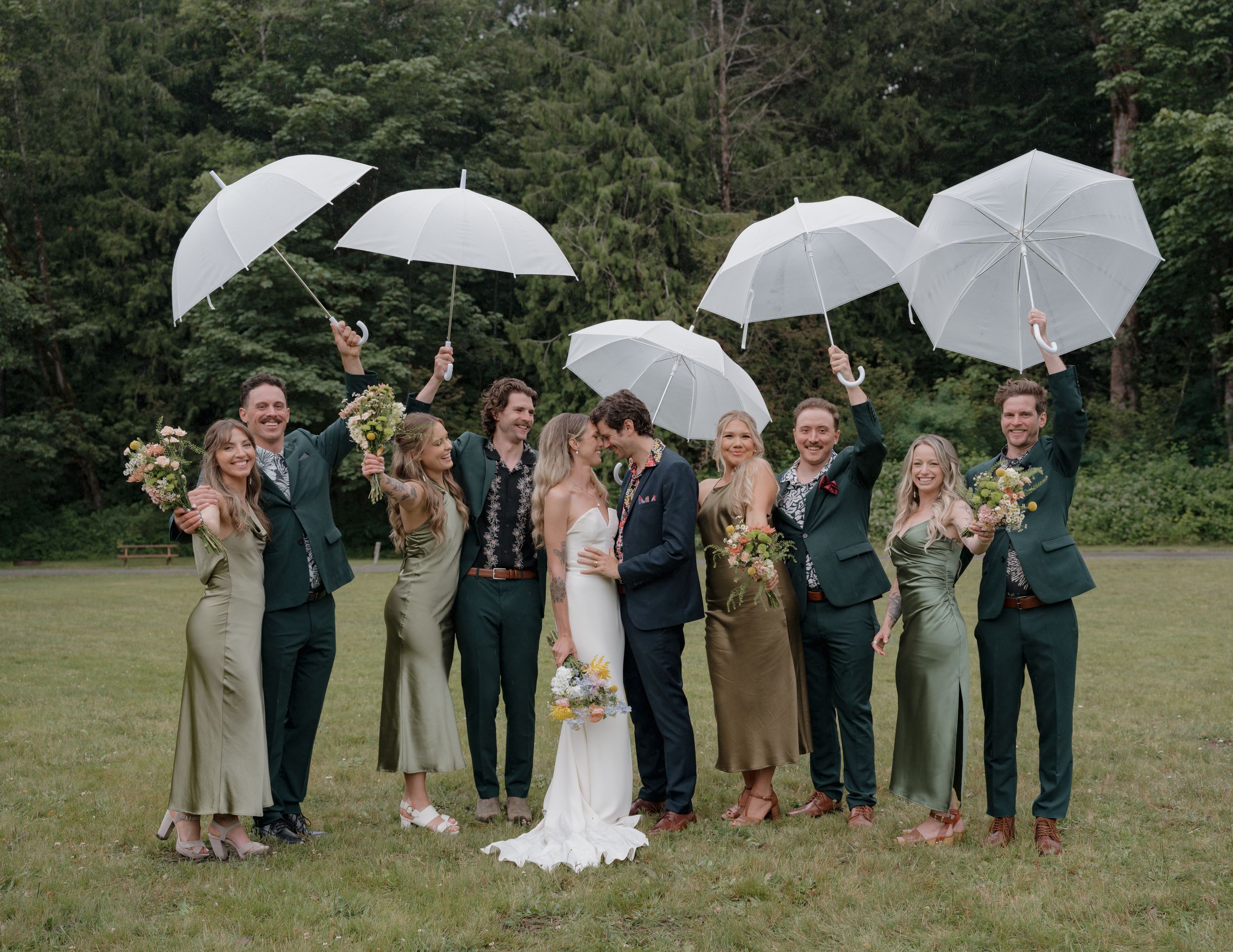 Wedding party stands in a line on grass holding white umbrellas while wearing formal attire and holding bouquets.