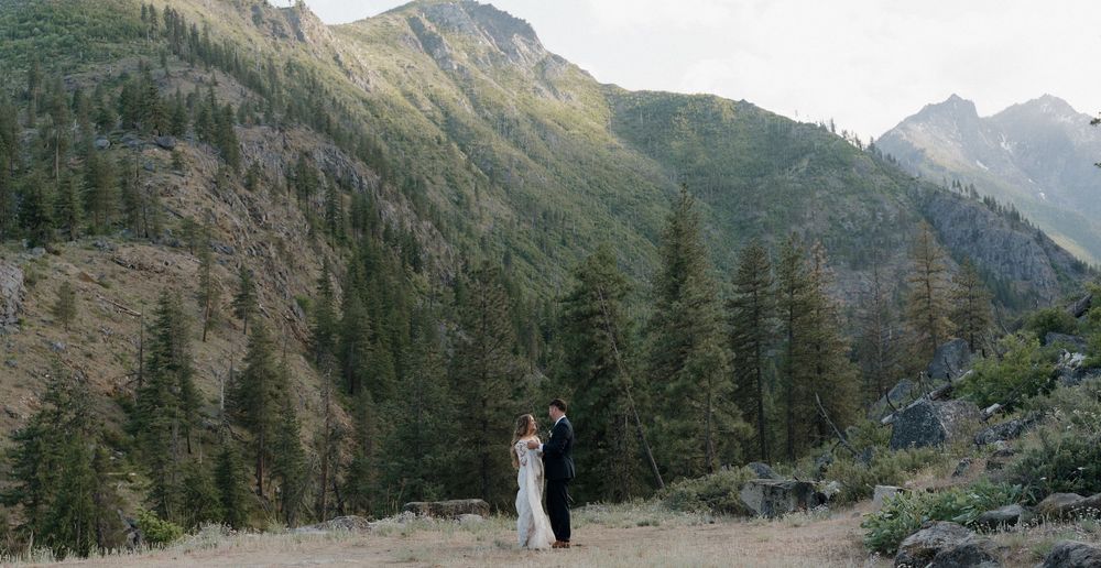A couple stands together in a scenic mountain valley surrounded by evergreen forest and rugged peaks.