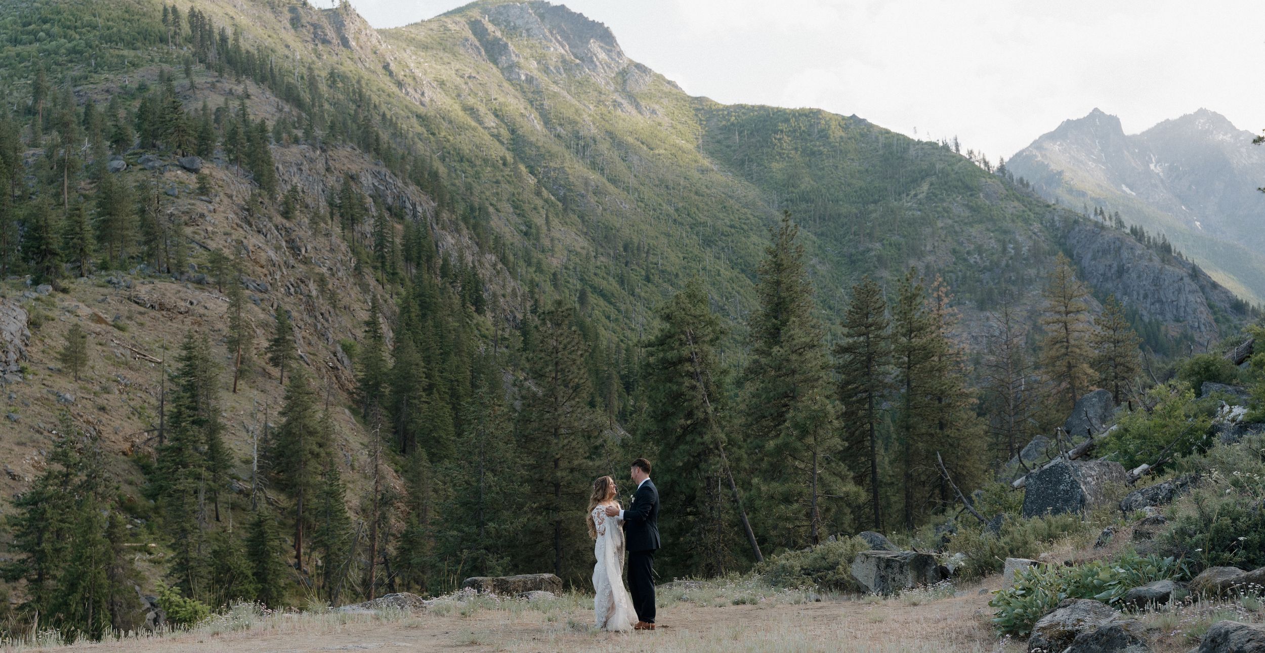 A couple stands together in a scenic mountain valley surrounded by evergreen forest and rugged peaks.