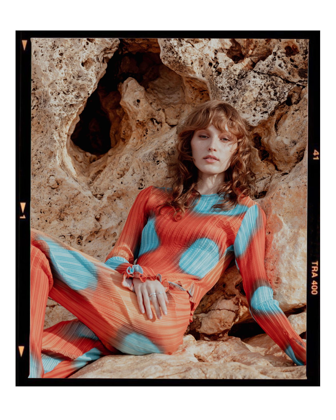 A fashion portrait featuring a vibrant coral and turquoise patterned outfit against textured rocks.