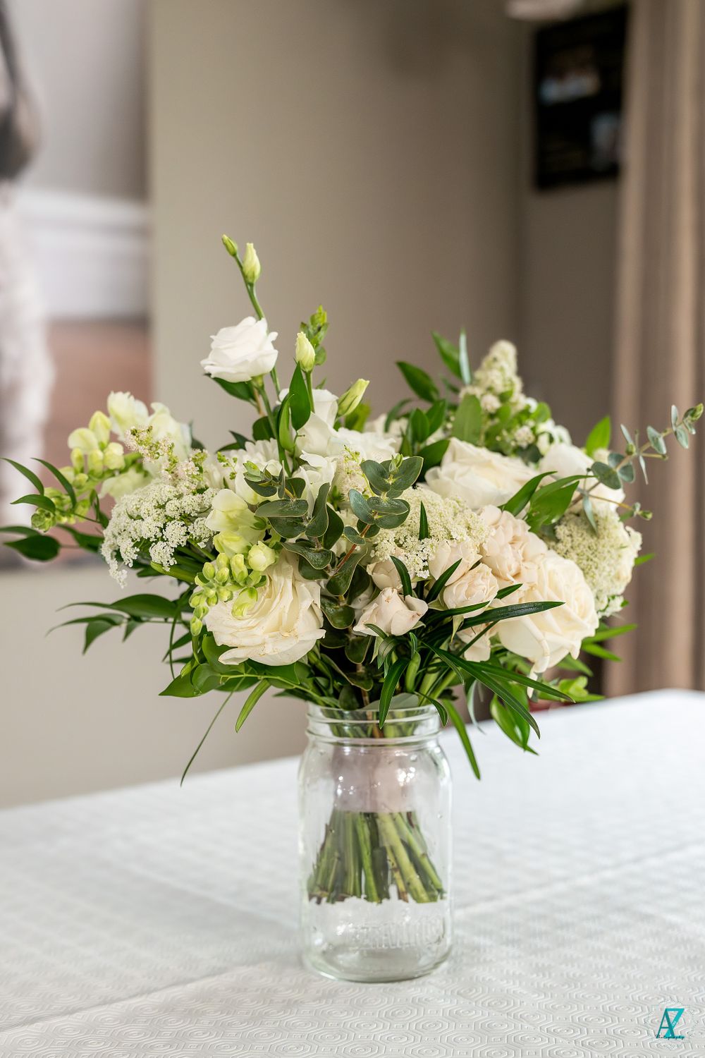 White and green floral arrangement with roses and mixed flowers in a clear mason jar vase.