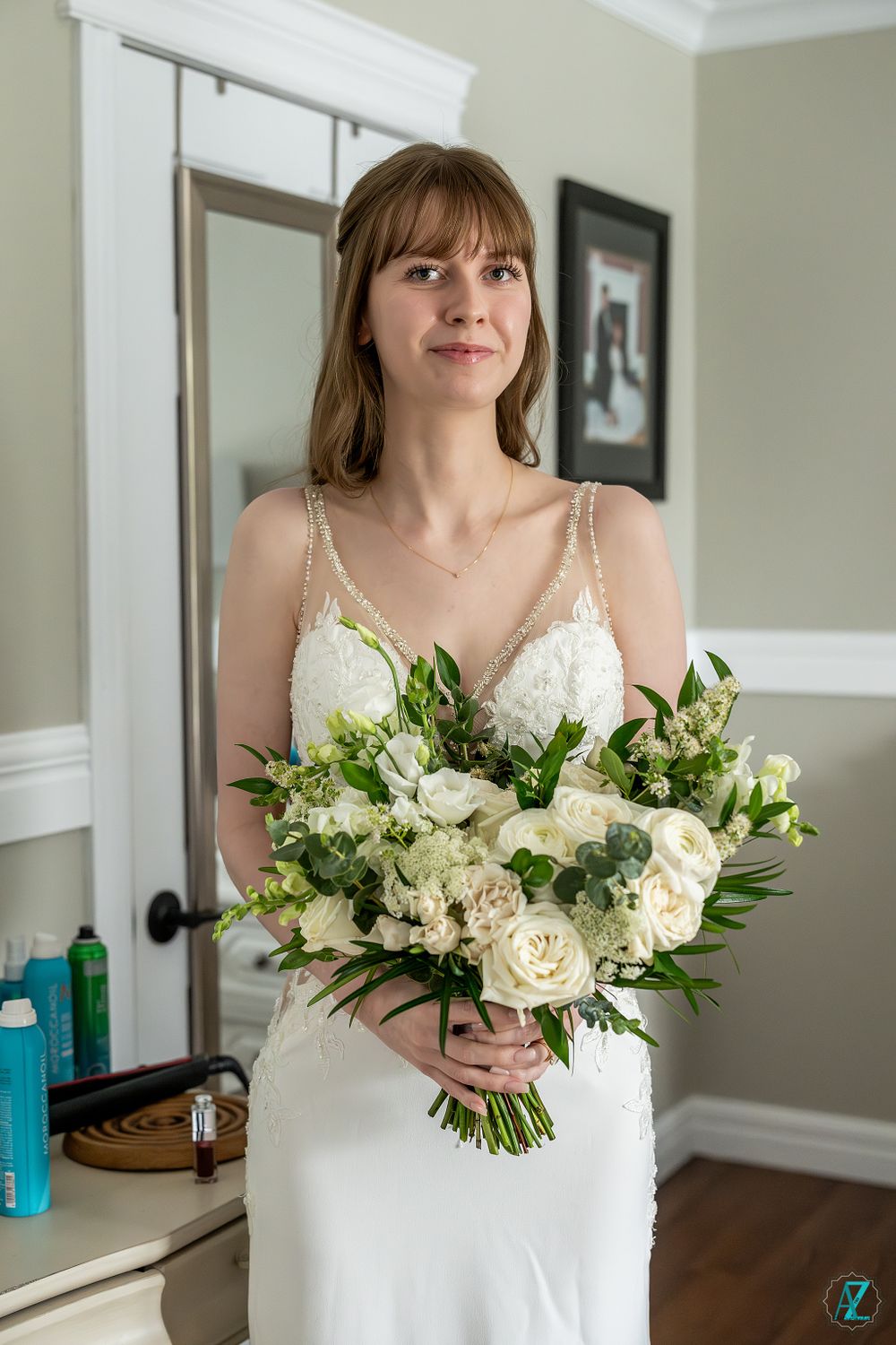 A bride holds a romantic bouquet of white roses and greenery while standing in a bridal suite.
