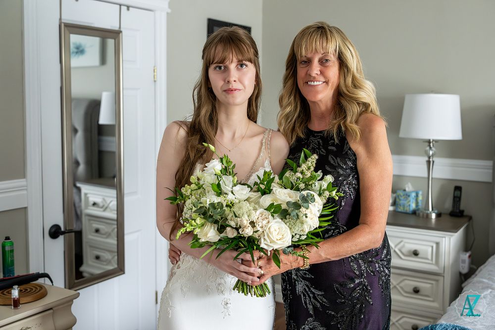 Two women in formal attire pose together, holding a wedding bouquet.
