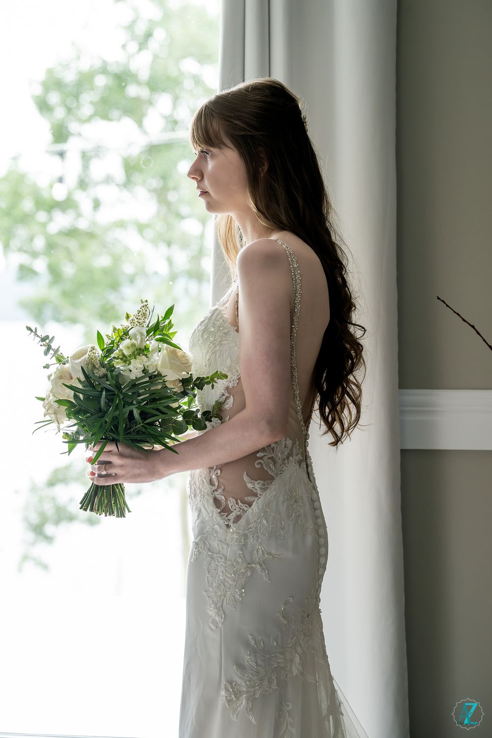 A side profile of a bride in lace gown holding a lush bouquet by a window.