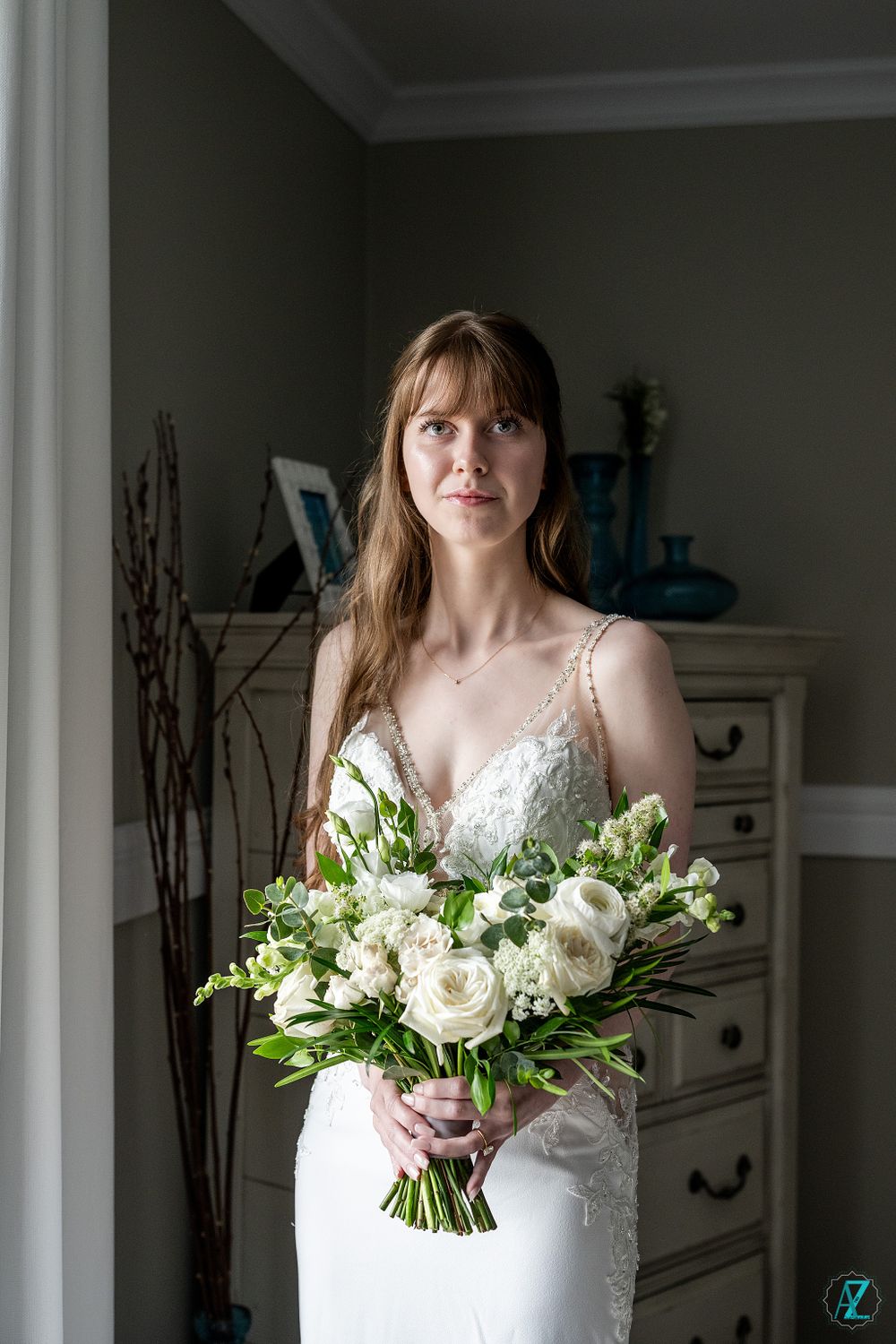 A bridal portrait by a window showcasing an elegant white gown and lush floral bouquet.