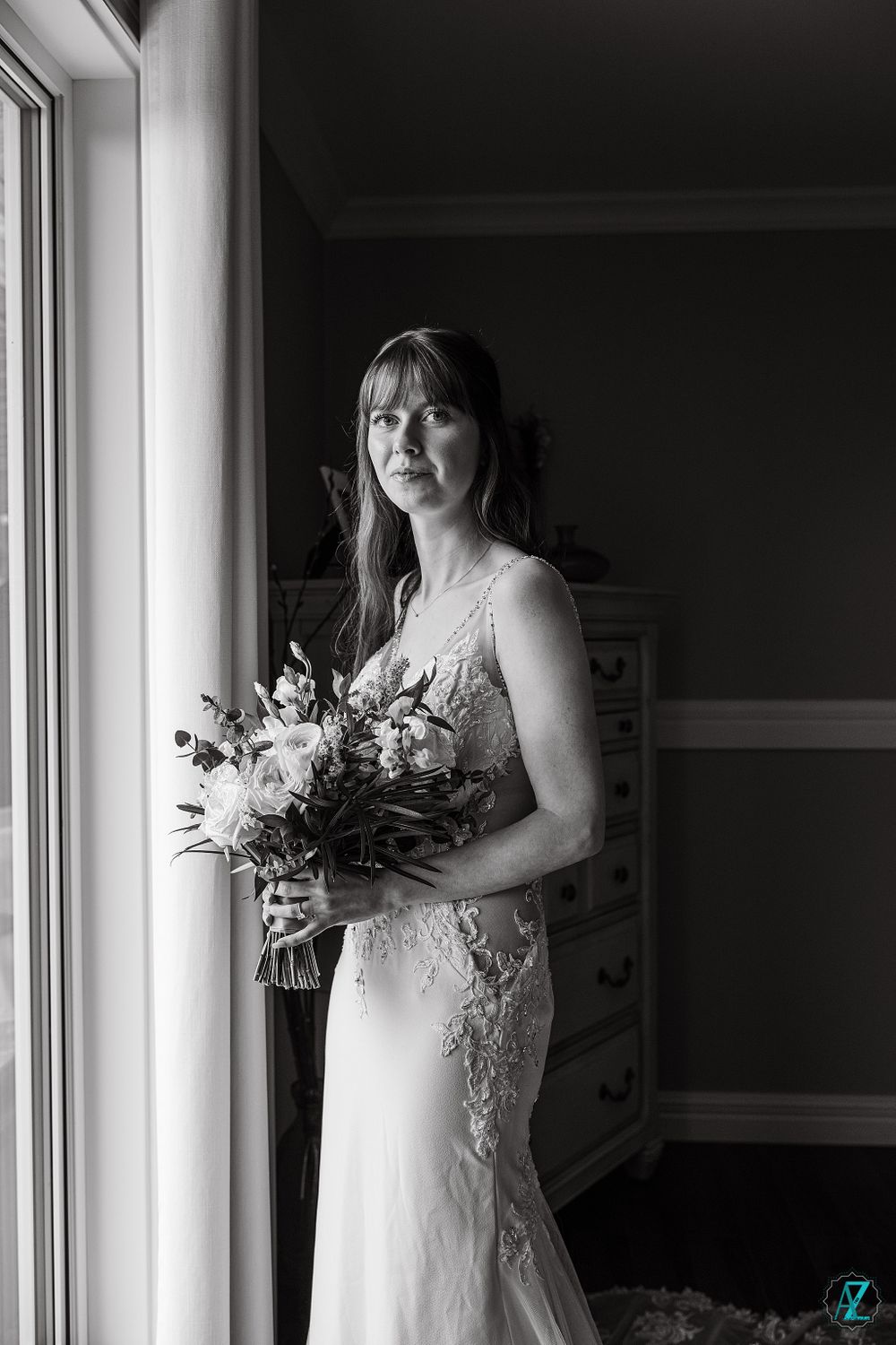 A black and white photo of a bride in a lace wedding dress holding a floral bouquet near a window.