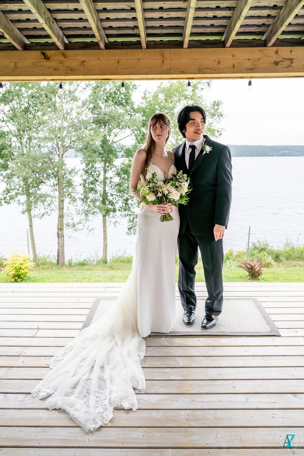A wedding couple poses on a lakeside deck with a beautiful flowing white dress and classic black suit.