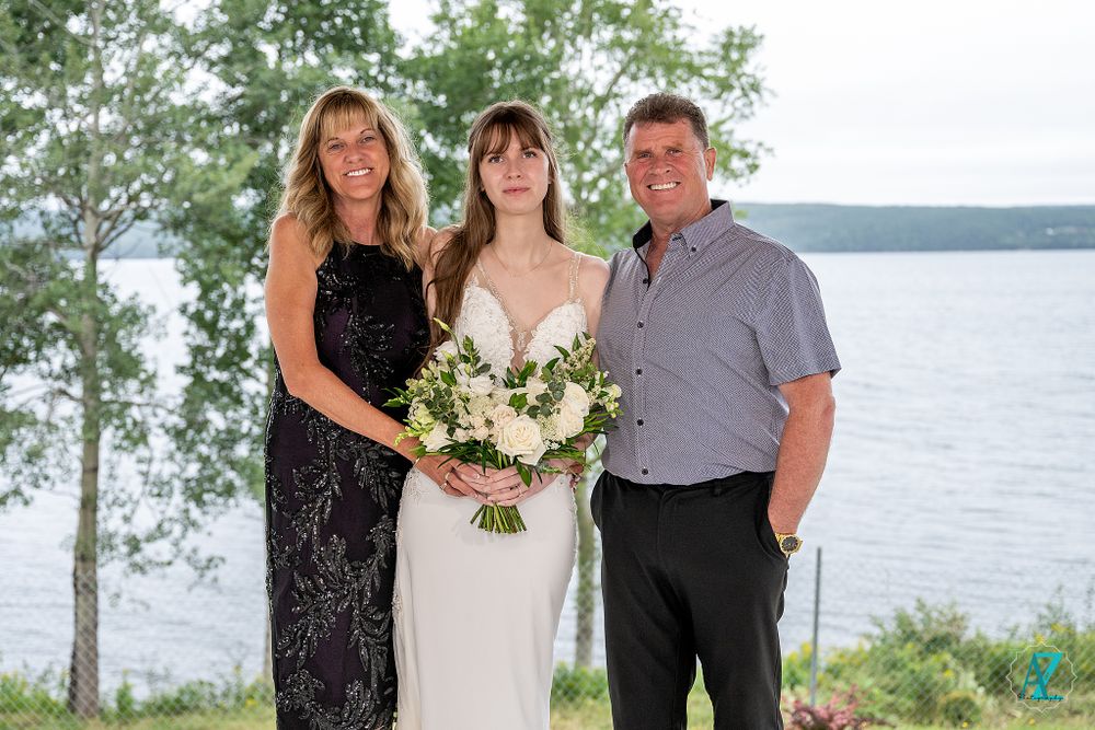 Three wedding guests pose together outdoors with lake and trees in background.
