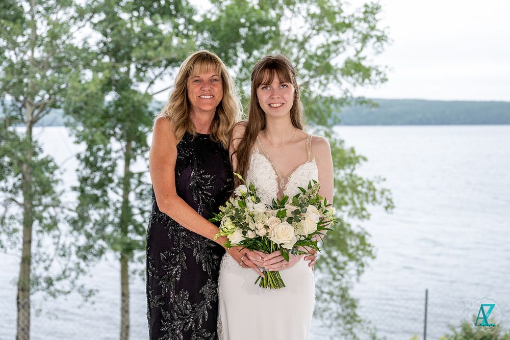 Two wedding attendees pose with floral bouquet against scenic lakeside background.